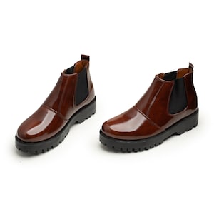 May include: A pair of brown Chelsea boots with a glossy finish. The boots have a black elastic side panel and a thick, black lug sole. The footwear is designed for everyday wear.