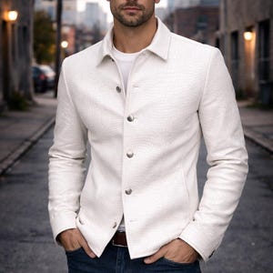 Könnte beinhalten: Eine weiße, strukturierte Jacke mit silbernen Knöpfen, kombiniert mit einer blauen Jeans. Die Jacke hat einen Kragen und ist zugeknöpft. Das Model steht in einer Gasse mit den Händen in den Taschen.