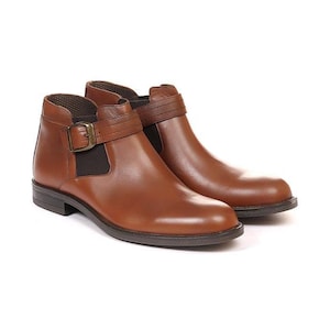 May include: A pair of brown leather ankle boots. The boots have a rounded toe, a strap with a buckle, and a dark brown sole. The leather has a polished finish. The boots are designed for everyday wear.