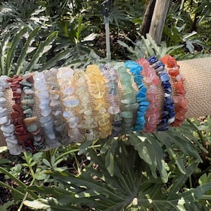 Op de afbeelding: Een verzameling edelsteen-chiparmbanden in verschillende kleuren, waaronder wit, rood, groen, blauw, roze en oranje. De armbanden worden getoond op een rol van bruin textiel tegen een achtergrond van groen gebladerte.