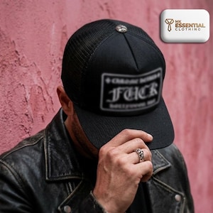 Puede incluir: Gorra de camionero negra con parte trasera de malla y visera curva. La gorra presenta un emblema plateado en la parte superior y un parche con la palabra "FUCK" en la parte delantera. La persona lleva una chaqueta de cuero negra.