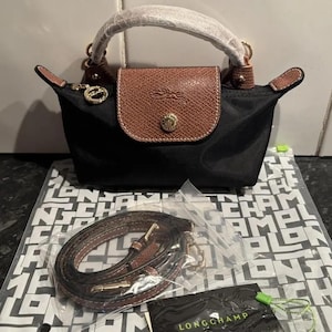 May include: A black nylon handbag with a brown leather flap featuring a gold-tone snap button and embossed logo. The bag has a top handle wrapped in protective paper, a zipper closure with a circular pull, and a detachable brown leather shoulder strap.