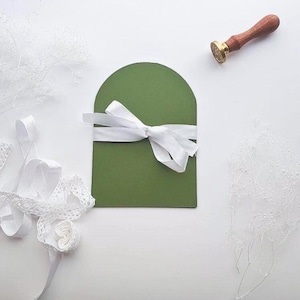 May include: An olive green card with a white ribbon tied in a bow. A wooden and gold wax seal stamp is in the upper right corner. White lace ribbon and decorative sprigs are in the image.
