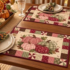 Puede incluir: Dos manteles individuales rectangulares acolchados con un estampado central de calabazas variadas en tonos rosa, verde y rojo oscuro, rodeados por un borde de patchwork de cuadrados en colores otoñales coordinados, incluyendo burdeos, salvia y crema.