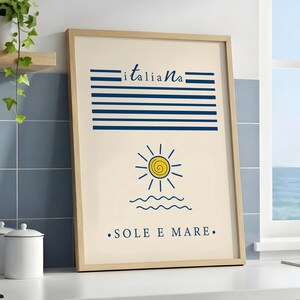 May include: A framed wall art print with a light beige background featuring the word "italiana" in blue script above horizontal blue lines. Below, a yellow sun with a spiral center sits above blue waves and the text "SOLE E MARE" in blue.