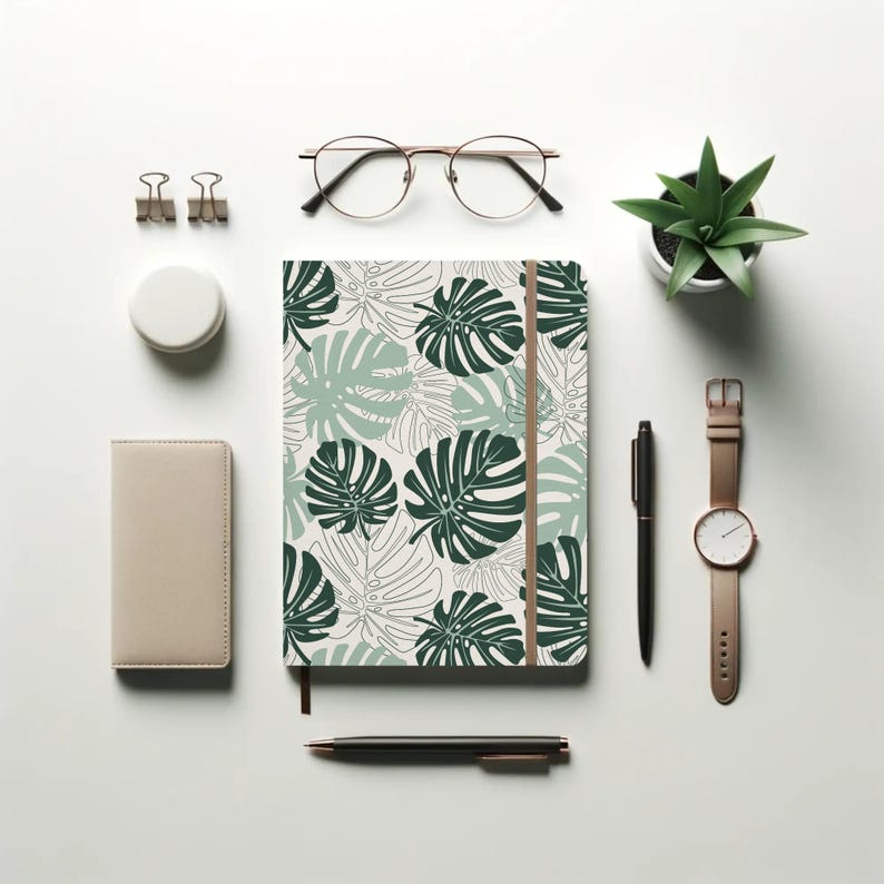 K&ouml;nnte beinhalten: Ein Notizbuch mit gr&uuml;nem Monstera-Blattmuster, ein ledergebundenes Notizbuch in Beige, eine kleine Topf-Sukkulente, eine Brille mit goldfarbenem Rahmen, eine Uhr und Stifte sind auf einer wei&szlig;en Oberfl&auml;che angeordnet.