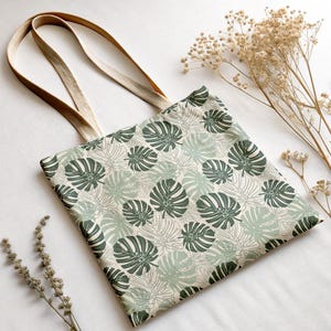 K&ouml;nnte beinhalten: Beige Tragetasche mit einem sich wiederholenden Muster aus gr&uuml;nen Monstera-Bl&auml;ttern. Die Tasche hat lange, beige Tr&auml;ger und besteht aus einem robusten Stoff. Der Hintergrund ist wei&szlig;, mit getrockneten Blumen.