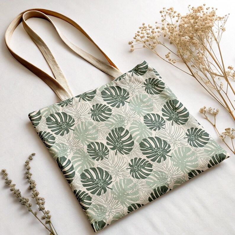 K&ouml;nnte beinhalten: Beige Tragetasche mit einem sich wiederholenden Muster aus gr&uuml;nen Monstera-Bl&auml;ttern. Die Tasche hat lange, beige Tr&auml;ger und besteht aus einem robusten Stoff. Der Hintergrund ist wei&szlig;, mit getrockneten Blumen.