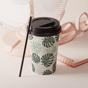 K&ouml;nnte beinhalten: Ein Einweg-Kaffeebecher mit schwarzem Deckel und schwarzem Strohhalm. Der Becher ist mit einem Muster aus gr&uuml;nen Monstera-Bl&auml;ttern auf cremefarbenem Hintergrund verziert. Ein rosa Ventilator und eine Brille sind im Hintergrund.