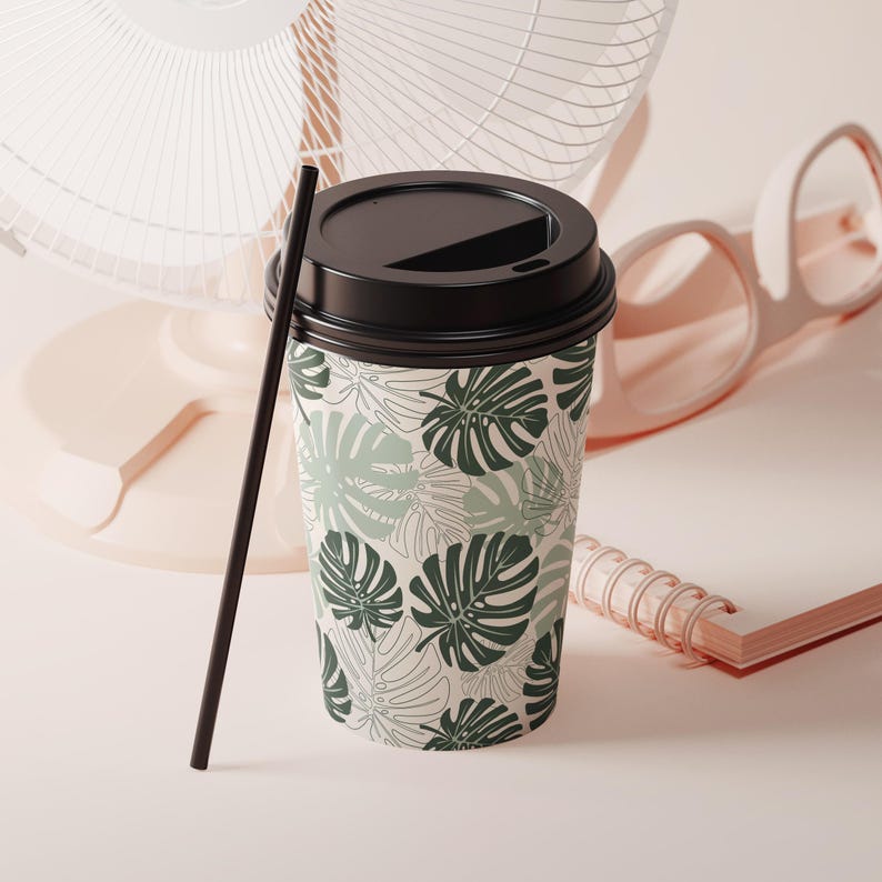 K&ouml;nnte beinhalten: Ein Einweg-Kaffeebecher mit schwarzem Deckel und schwarzem Strohhalm. Der Becher ist mit einem Muster aus gr&uuml;nen Monstera-Bl&auml;ttern auf cremefarbenem Hintergrund verziert. Ein rosa Ventilator und eine Brille sind im Hintergrund.