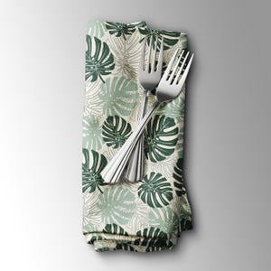 K&ouml;nnte beinhalten: Eine gefaltete Stoffserviette mit einem tropischen Bl&auml;ttermuster in Gr&uuml;n- und Wei&szlig;t&ouml;nen, mit zwei silbernen Gabeln darauf. Das Design der Serviette zeigt gro&szlig;e, stilisierte Monstera-Bl&auml;tter.