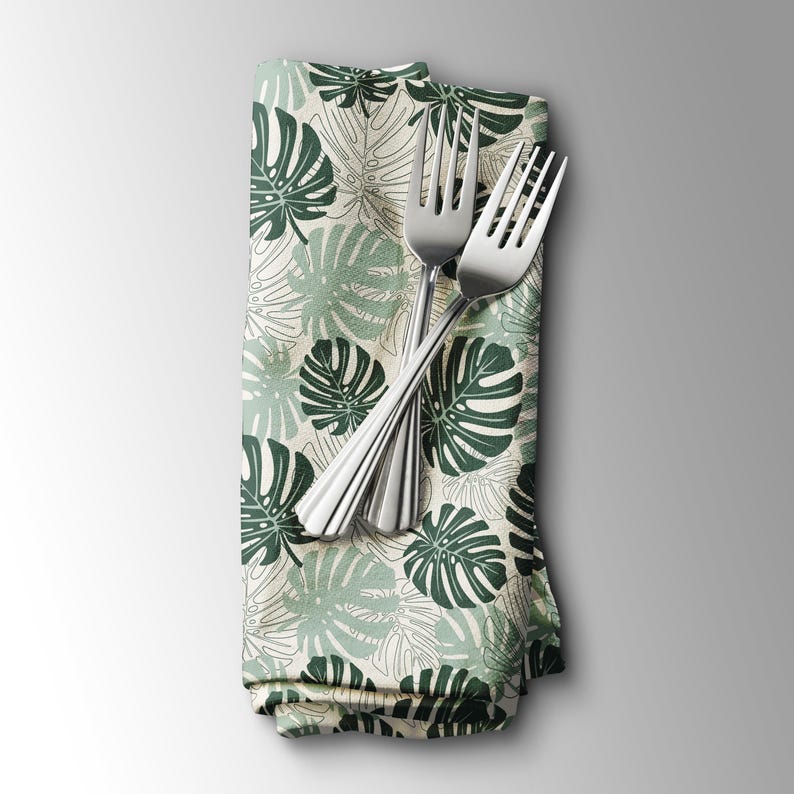 K&ouml;nnte beinhalten: Eine gefaltete Stoffserviette mit einem tropischen Bl&auml;ttermuster in Gr&uuml;n- und Wei&szlig;t&ouml;nen, mit zwei silbernen Gabeln darauf. Das Design der Serviette zeigt gro&szlig;e, stilisierte Monstera-Bl&auml;tter.