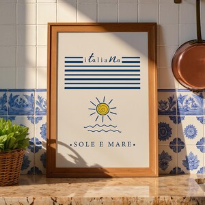 May include: A framed rectangular wall art print featuring a cream background with horizontal dark blue stripes, the word "italiana" in blue script, a yellow sun icon, wavy blue lines, and the text "SOLE E MARE" in blue capital letters.