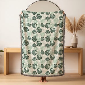 K&ouml;nnte beinhalten: Eine gewebte Wurkdecke mit einem sich wiederholenden Muster aus gr&uuml;nen Monstera-Bl&auml;ttern auf cremefarbenem Hintergrund. Die Decke hat einen Fransenrand und wird vor einer hellen Wand und einem Holztisch pr&auml;sentiert.
