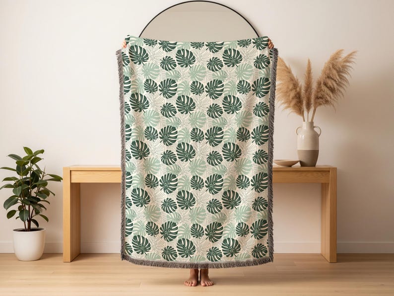 K&ouml;nnte beinhalten: Eine gewebte Wurkdecke mit einem sich wiederholenden Muster aus gr&uuml;nen Monstera-Bl&auml;ttern auf cremefarbenem Hintergrund. Die Decke hat einen Fransenrand und wird vor einer hellen Wand und einem Holztisch pr&auml;sentiert.