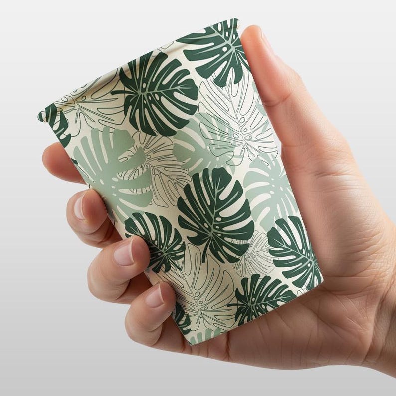 K&ouml;nnte beinhalten: Ein Pappbecher mit einem Muster aus gr&uuml;nen und wei&szlig;en tropischen Bl&auml;ttern. Der Becher wird in einer Hand gehalten, wodurch das Design zur Geltung kommt. Die Bl&auml;tter sind eine Mischung aus Voll- und Linienstilen, wodurch ein geschichteter Effekt entsteht.