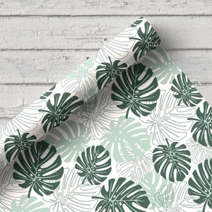 K&ouml;nnte beinhalten: Gerolltes Geschenkpapier mit einem sich wiederholenden Muster aus dunkelgr&uuml;nen und hellgr&uuml;nen Monstera-Bl&auml;ttern auf wei&szlig;em Hintergrund. Die Bl&auml;tter haben detaillierte Aderstrukturen. Das Papier ist gerollt und zeigt das Design.
