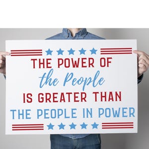 Peut inclure: Une affiche blanche avec des rayures rouges et bleues et des étoiles. Le texte sur l'affiche est "THE POWER OF the People IS GREATER THAN THE PEOPLE IN POWER".
