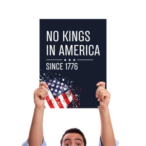 May include: A person holding a navy blue poster with the text "NO KINGS IN AMERICA SINCE 1776" in white. The bottom of the poster features a distressed American flag design with red and white stripes and white stars.