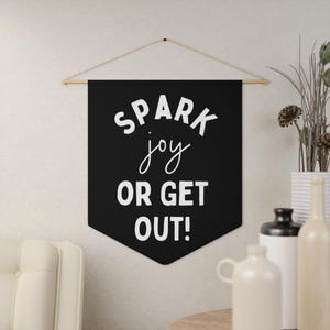以下が含まれることがあります： 白い文字で「SPARK joy OR GET OUT！」と書かれた黒い壁掛け。バナーは、天然のコードが付いた木の棒から吊り下げられています。背景には装飾品が含まれています。