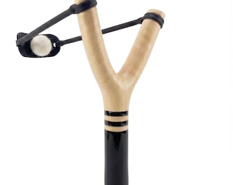 May include: A wooden slingshot with a light brown Y-shaped frame. The handle is painted black with two black stripes. A black rubber band and pouch hold a white projectile, ready to launch. The slingshot is designed for recreational use.