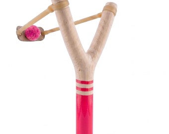 May include: A wooden slingshot with a pink and white handle and a pink ball. The slingshot is made of light-colored wood with a Y-shaped frame. The handle is painted pink with three white stripes.