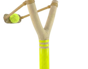 May include: A wooden slingshot with a neon yellow handle and three yellow stripes. The slingshot has a light brown rubber band and is holding a yellow tennis ball. The slingshot is on a white background.