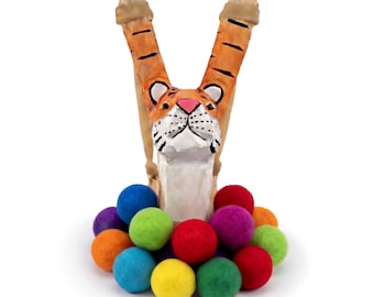 May include: A whimsical wooden tiger-shaped sculpture with orange and black stripes, featuring a white face and pink nose. The tiger holds two upright wooden pieces. A colorful array of felt balls in various colors surrounds the base.