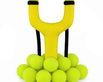 May include: A bright yellow slingshot with black bands and accents, resting on a bed of neon green tennis balls. The slingshot has a classic Y-shape design with black bands for launching projectiles. The tennis balls are arranged in a circular pattern.