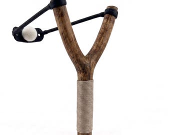 May include: A wooden slingshot with a Y-shaped frame. The slingshot has a brown wooden handle wrapped with tan twine. The black rubber band holds a white felt ball. The slingshot is set against a white background.