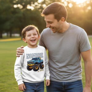 Puede incluir: Una sudadera blanca con la palabra "NOMBRE" en letras azul claro. Debajo, un camión monstruo azul con ruedas amarillas y el texto "Race to Victory". La imagen muestra a un niño y un adulto sonriendo.