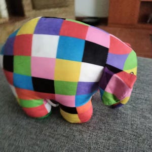 May include: A plush elephant toy with a patchwork design in vibrant colors including red, yellow, blue, green, pink, and black. The toy has a trunk, ears, and four legs. The elephant is sitting on a gray surface.
