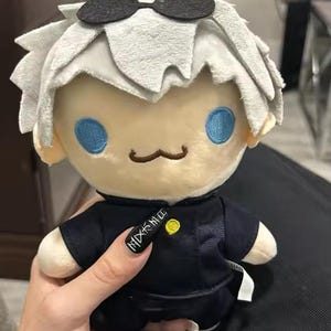 May include: A plush doll of a character with white hair, blue eyes, and a small mustache. The doll wears a black outfit and sunglasses. The doll is held by a person.