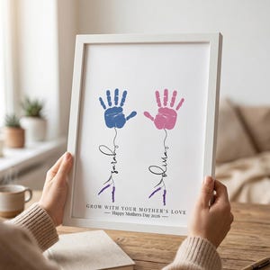Könnte beinhalten: Gerahmter Druck mit blauen und rosa Handabdrücken, den Namen "Sarah" und "Olivia" in Schreibschrift und Lavendelzweigen. Der Text lautet: "Grow with your Mother's Love - Happy Mothers Day 2026". Ein liebevolles Geschenk.