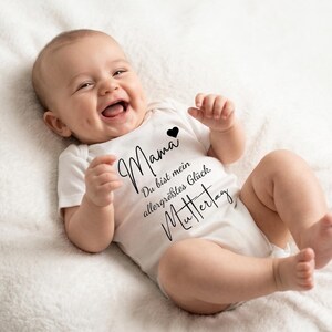 Könnte beinhalten: Ein lächelndes Baby in einem weißen Body mit schwarzer Schrift, die "Mama, Du bist mein allergrößtes Glück, Muttertag" und ein kleines schwarzes Herz zeigt. Das Baby liegt auf einer weißen, strukturierten Oberfläche.