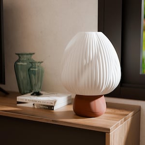 May include: A white, textured table lamp with a rounded top and a terracotta-coloured base. The lamp is on a wooden surface, with two green glass vases and a book titled "THE FASHION BOOK" in the background. The lamp's design is reminiscent of a mushroom.