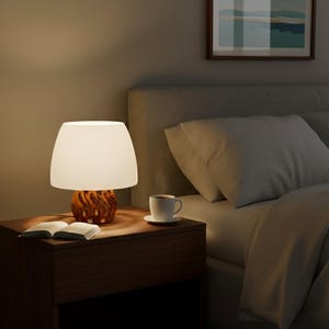 Op de afbeelding: Een tafellamp met een witte lampenkap en een bruine, getextureerde voet. De lamp staat op een donkerhouten nachtkastje naast een bed met wit beddengoed. Een kopje koffie en een open boek staan ook op het nachtkastje.