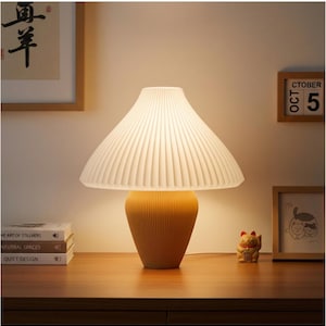 May include: A table lamp featuring a matte mustard yellow base with vertical ribbing and a wide, white, pleated conical shade. The lamp emits a warm, soft glow, illuminating the surrounding area with a gentle light while resting on a flat surface.
