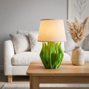 May include: A contemporary table lamp featuring a green, abstract base and a beige lampshade. The base has a distinctive, organic form. The lamp is placed on a wooden table, with a white sofa in the background. It is a decorative home decor item.