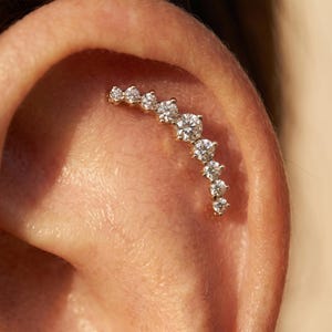 Peut inclure: Une boucle d'oreille grimpante en or composée d'une rangée incurvée de huit pierres claires rondes de tailles graduées, la plus grande étant située près du centre de l'arc, épousant le contour naturel du cartilage de l'oreille.