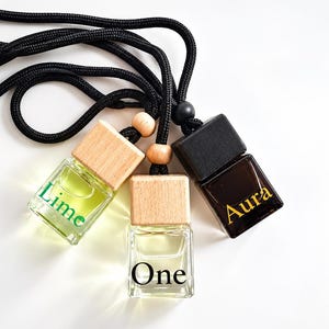 Könnte beinhalten: Drei kleine quadratische Glasflaschen mit Holzverschlüssen, die an einer schwarzen geflochtenen Kordel hängen. Die linke Flasche enthält eine hellgrüne Flüssigkeit und trägt die Aufschrift Lime. Die mittlere Flasche ist klar und trägt die Aufschrift One.