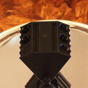 May include: A black plastic pencil sharpener with a faceted, diamond-like shape and multiple circular openings along the top edge, positioned centrally on a reflective silver circular tray.