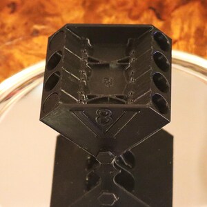 May include: A black plastic V8 engine block model featuring a prominent number 8 embossed on the front face. The object has a rectangular top section with circular openings along the sides and a solid base, displaying a detailed mechanical design.
