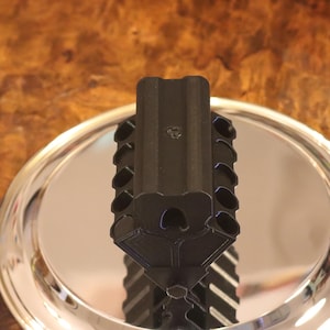 May include: A matte black, V-shaped engine block model featuring a series of cylindrical ports along its sides, positioned vertically atop a circular, polished silver-toned metallic base that reflects the object's structure.