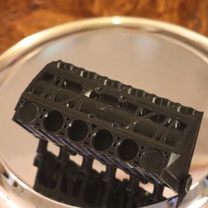 Könnte beinhalten: Ein schwarzes, 3D-gedrucktes Modell eines Verbrennungsmotorblocks mit einer Reihe kreisförmiger Zylinderbohrungen auf der Oberseite und einem detaillierten, strukturierten Rahmen mit verschiedenen Öffnungen und Stützrippen.