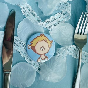 May include: A button with a cartoon illustration of a person with blonde hair and a blue background, placed on a light blue surface with a knife, fork, white lace ribbon, and light blue petals.