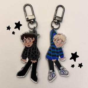 May include: Two acrylic keyrings with cartoon illustrations of young people. One shows a person in a black and grey camouflage hoodie. The other depicts a person in a blue and black striped sweater. Both keyrings have silver clasps and are decorated with black star shapes.