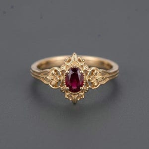 Puede incluir: Un anillo de oro con una piedra preciosa de color rojo intenso de corte ovalado en el centro. La banda presenta un intrincado trabajo de metal, incluyendo pequeños motivos florales a cada lado de la piedra y adornos en espiral alrededor del engaste.