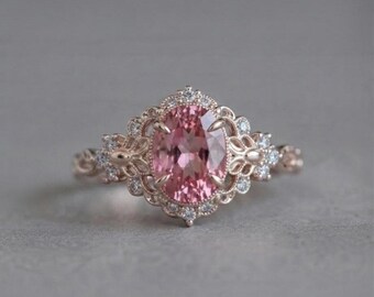 May include: A rose gold ring featuring a central oval-cut pink gemstone held by four prongs. The setting is surrounded by a halo of small round white stones and intricate metal filigree work that extends along the band of the ring.