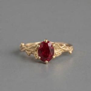 Könnte beinhalten: Ein goldener Ring mit einem zentralen, oval geschliffenen, tiefroten Edelstein, der von vier Krappen gehalten wird. Die Ringschiene ist mit einem strukturierten, erhabenen Blatt- und Rankenmuster verziert, das sich beidseitig erstreckt.
